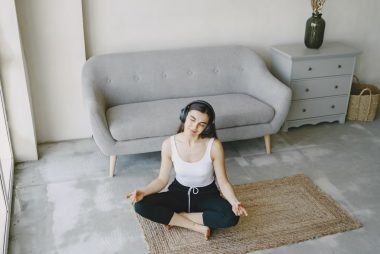 A young woman sits on a mat in a contemporary living room, meditating with headphones on.