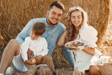 Smiling family of three enjoying a sunny day picnic with pastries on a hay field.